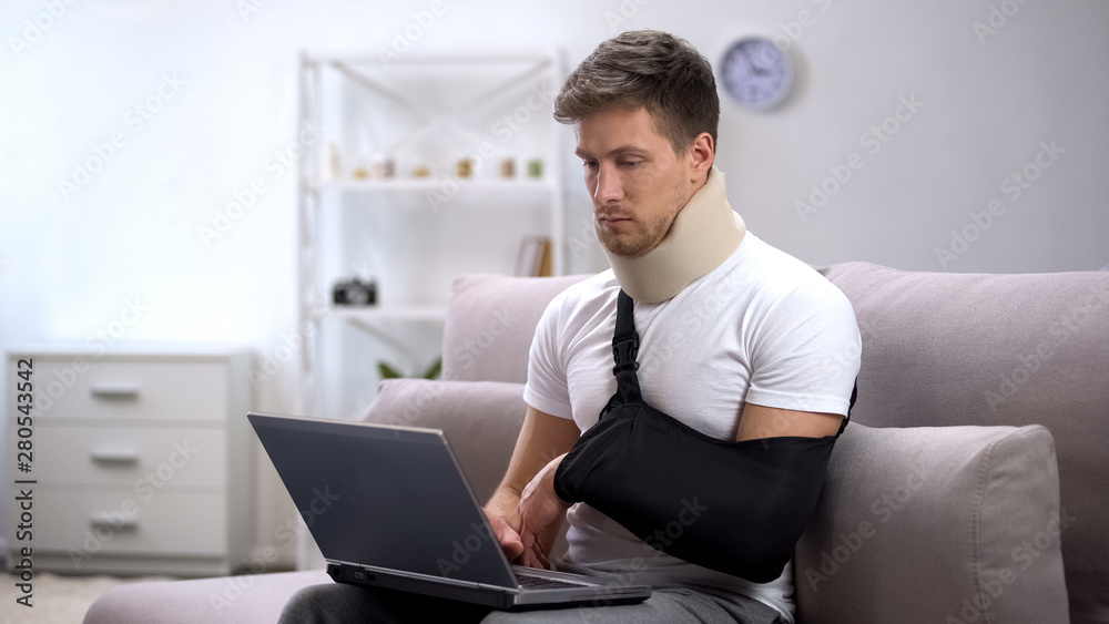 Male in foam cervical collar and arm sling typing one hand on laptop, freelance