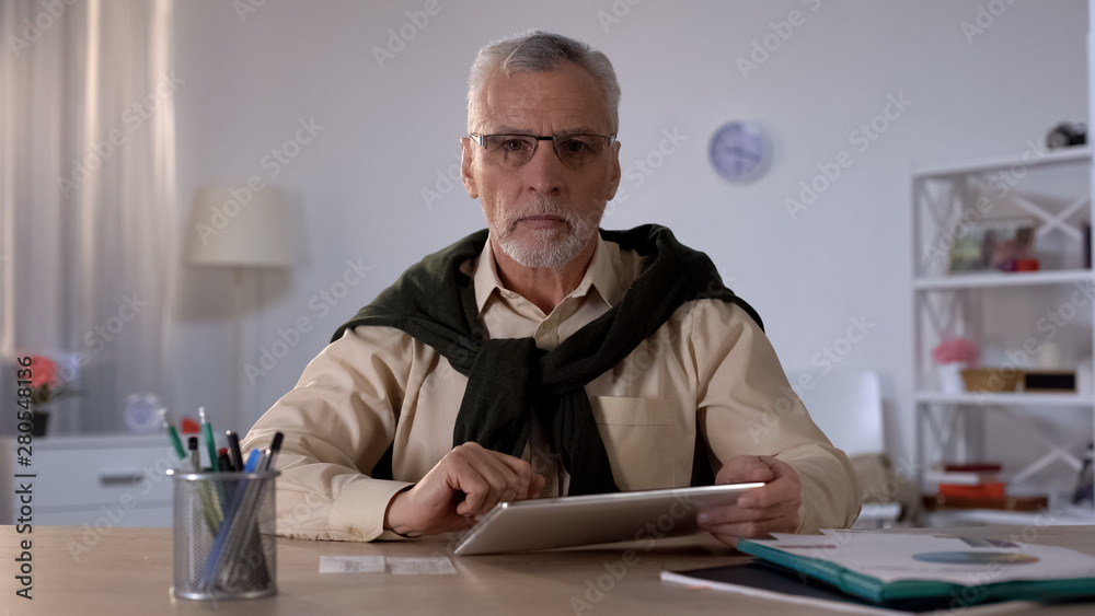 Serious senior man holding tablet and looking at camera, planning budget app