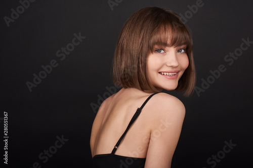 Beauty portrait of a beautiful young smiling girl with beautiful glowing skin and natural makeup on a black background in a black dress.