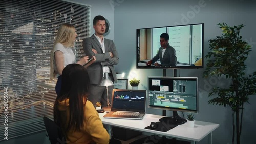 Mixed race team of video editors listening to chief commenting video watched on big screen. There is also personal computer with two displays in office.