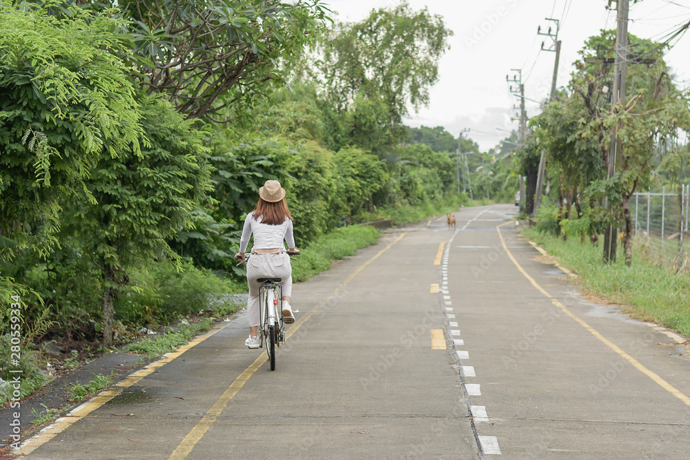 Fototapeta premium Pretty girl with straw hat is happy riding with bike down wide beautiful park alley with trees around on sunny summer day.