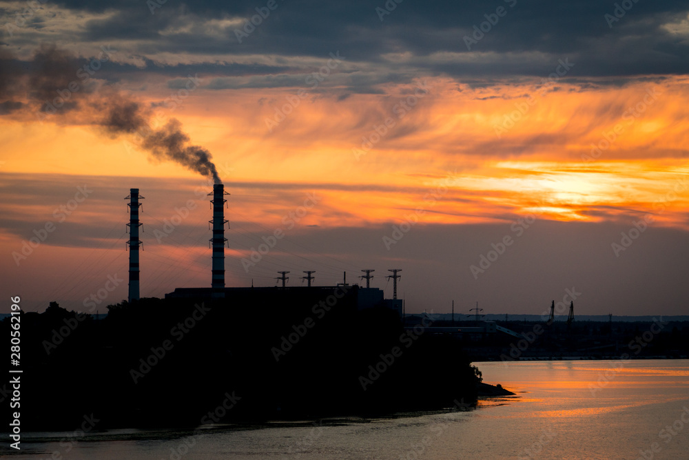 Sunset silhouette of industrial plant with a smoking pipe located on a river.