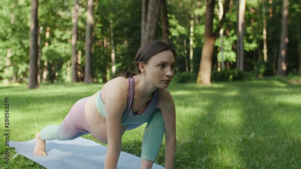 Pregnant woman doing yoga in nature outdoors on sunny day. Young expectant mother practice stretching yoga in in low lunge position on mat in green park with tall trees on background Pregnant woman doing yoga in nature outdoors on sunny day. Young expectant mother practice stretching yoga in in low lunge position on mat in green park with tall trees on background