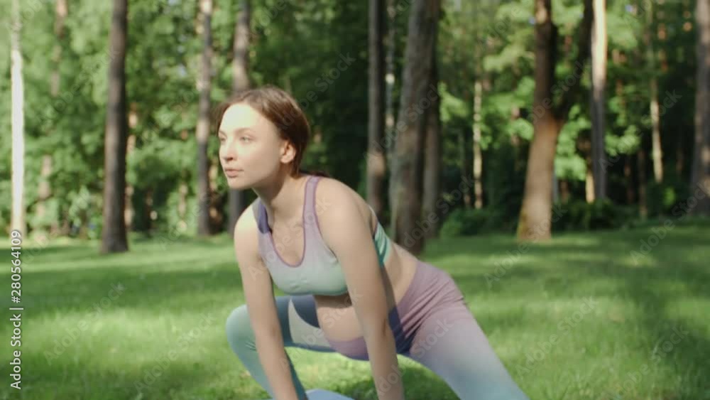 Pregnant woman doing yoga in nature outdoors on sunny day. Young future mother practice stretching yoga in in low lunge position on mat in green park with tall trees on background. Handheld camera Pregnant woman doing yoga in nature outdoors on sunny day. Young future mother practice stretching yoga in in low lunge position on mat in green park with tall trees on background. Handheld camera
