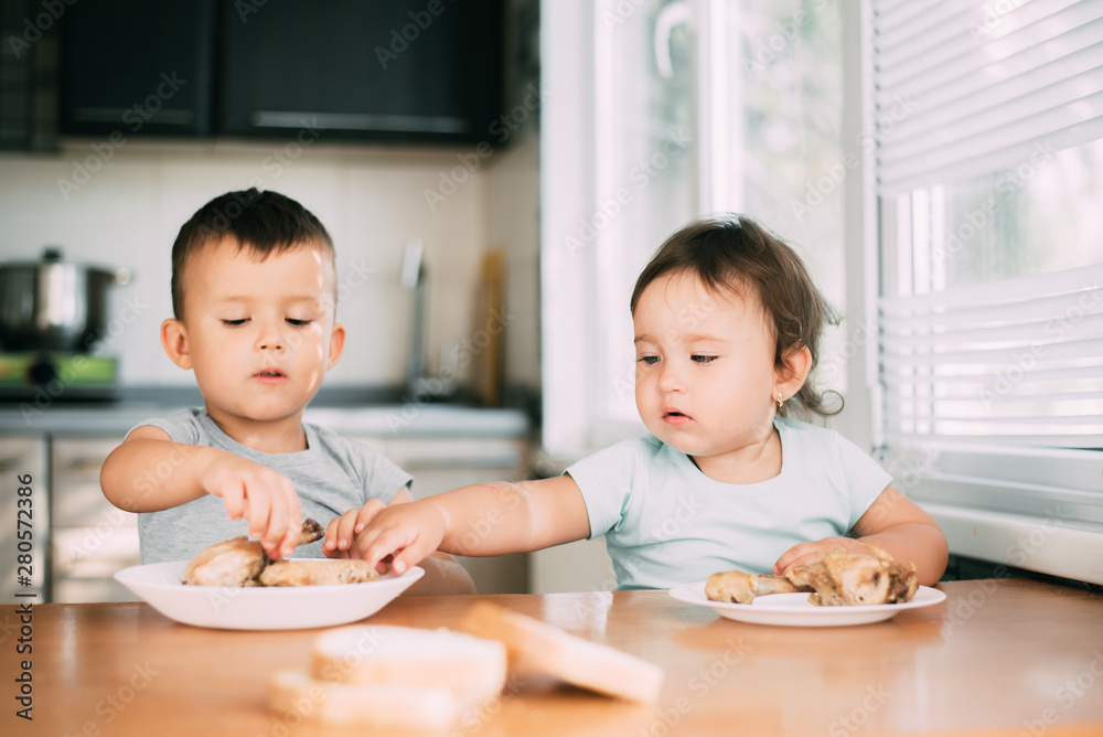 Kids boy and girl , brother and sister in the kitchen having fun and eating chicken with appetite Kids boy and girl , brother and sister in the kitchen having fun and eating chicken with appetite