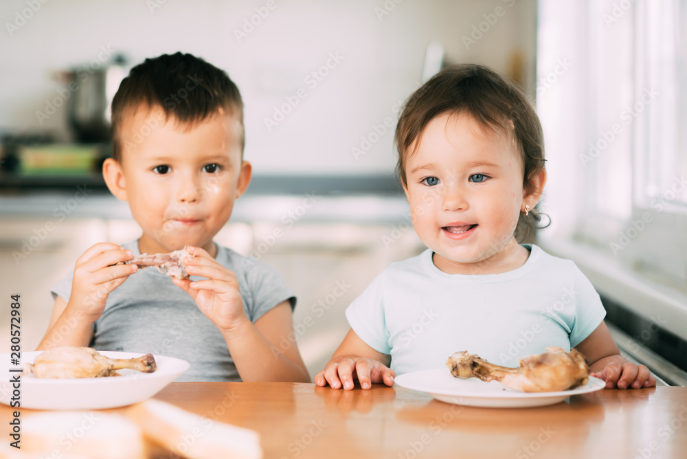 Kids boy and girl , brother and sister in the kitchen having fun and eating chicken with appetite Kids boy and girl , brother and sister in the kitchen having fun and eating chicken with appetite