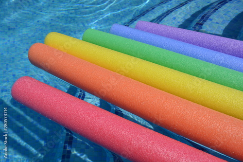 Rainbow coloured pool noodles floating in a swimming pool, fun summer vibes