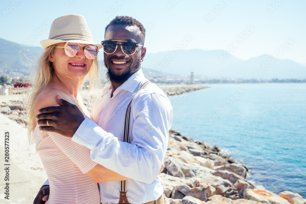 smiling happy european woman in straw hat and sexy pink dress walking with afro american ethnic man by summer beach with rock view in Turkey tropical resort
