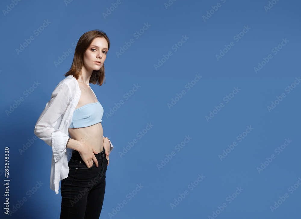 Portrait of the young modern woman. The serious beautiful thin brunette with equal hair in youth clothes. Blue background Portrait of the young modern woman. The serious beautiful thin brunette with equal hair in youth clothes. Blue background