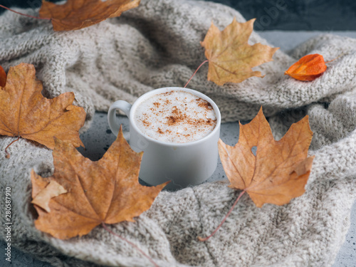 White Cup of latte coffee on gray table with a knitted scarf and a dry autumn maple leaves. Home atmosphere and comfort in cold weather