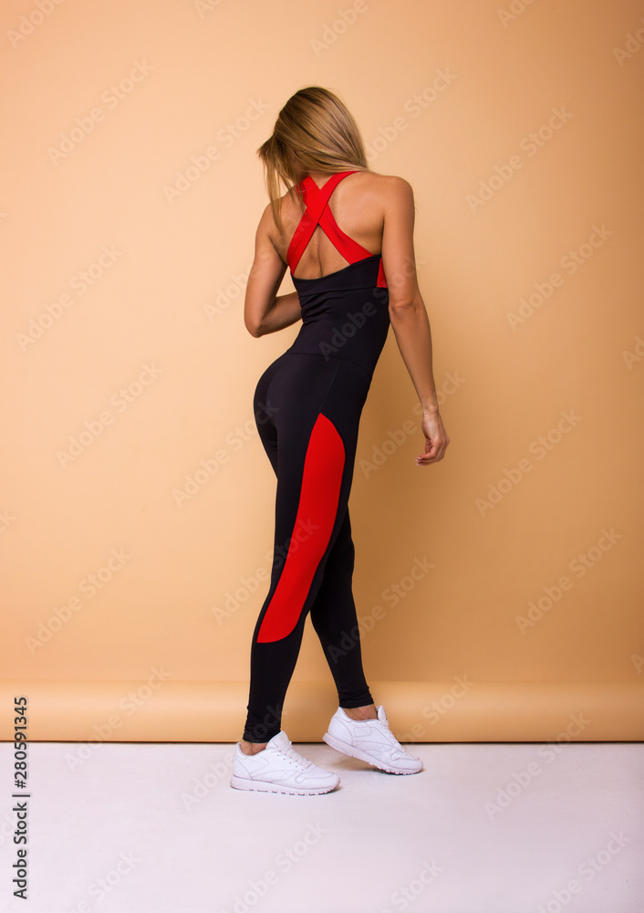 Obraz premium young sexy blonde girl in black costume with red stripes and white sneakers is standing from the back and looking down on the beige wall background, sport concept, free space