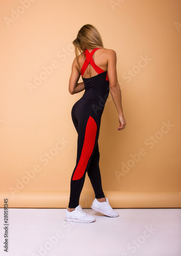 young sexy blonde girl in black costume with red stripes and white sneakers is standing from the back and looking down on the beige wall background, sport concept, free space