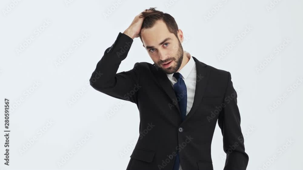 Attractive young businessman in black suit and blue tie smiling and fixing his hair while looking at the camera over gray background isolated Attractive young businessman in black suit and blue tie smiling and fixing his hair while looking at the camera over gray background isolated