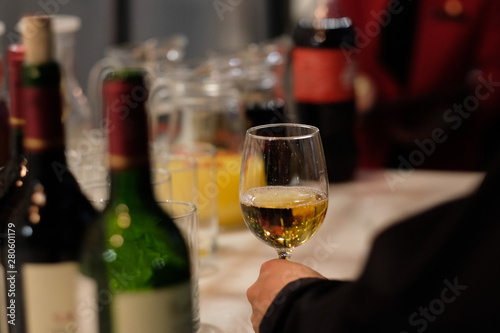 Over shoulder angle of one man‘s hand taking white wine glass with defocused bottles and glasses around. Drinking party concept 