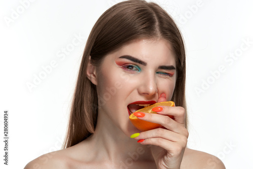 Closeup portrait of a beautiful young woman with bright color make-up biting half of an orange near the face.
