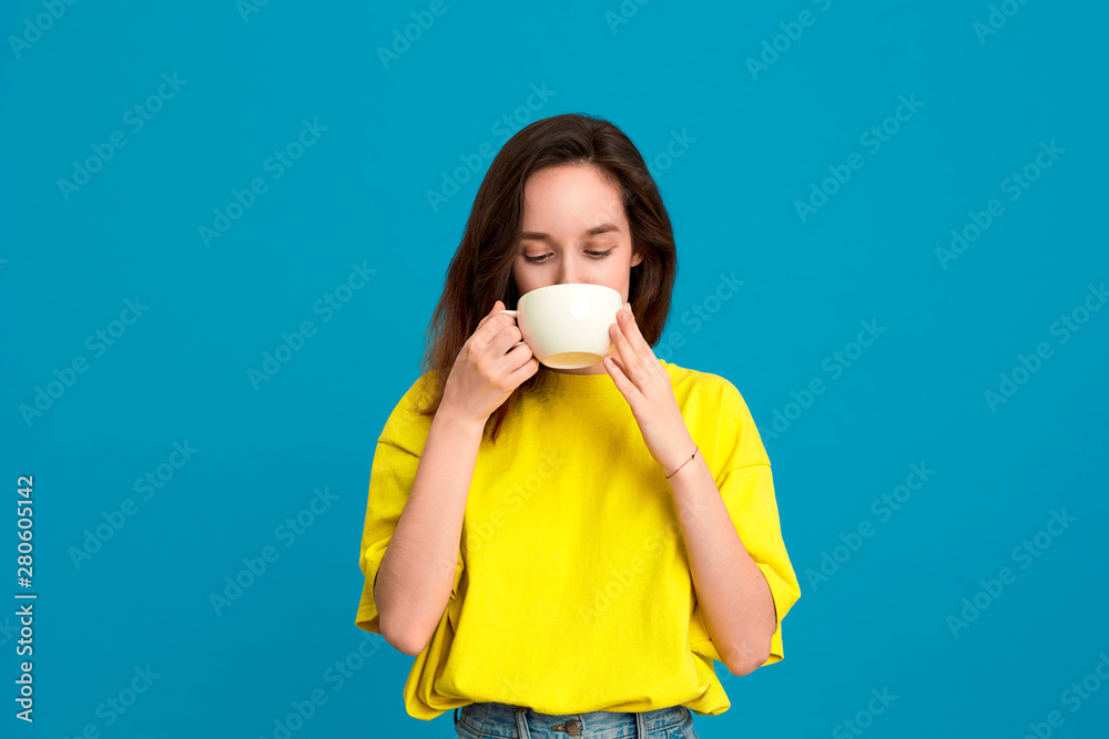 Pretty brunette woman in a yellow t-shirt drinking beverage from a white cup standing over blue background. Pretty brunette woman in a yellow t-shirt drinking beverage from a white cup standing over blue background.