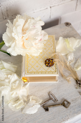 A beautiful white peony lies on a book with Golden pages. Near are delicate petals, vintage keys and embellishment on the neck. Vintage background in light white and beige.