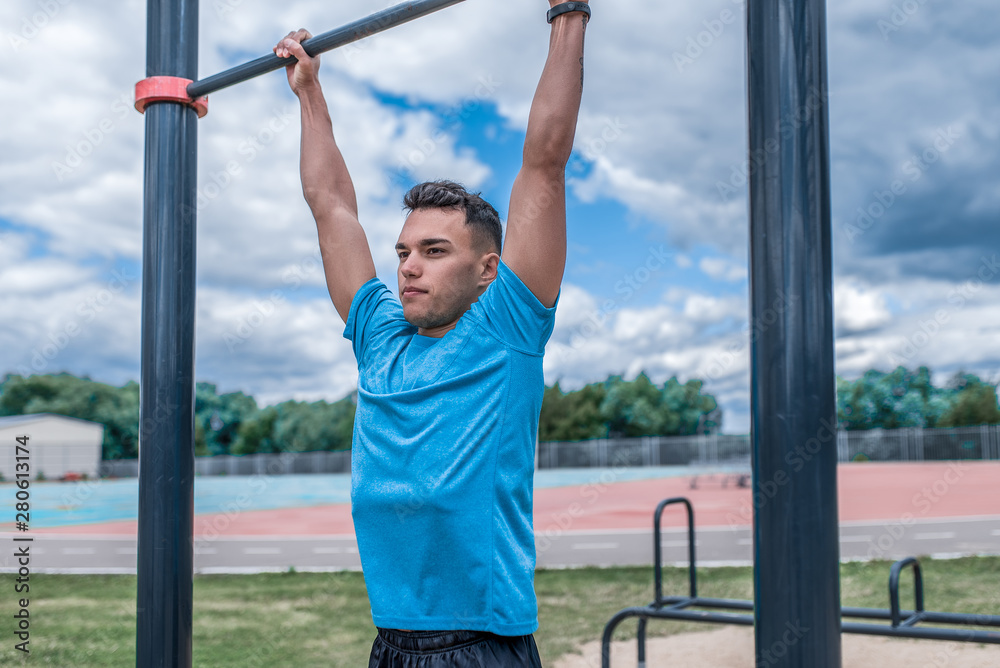 young guy, man sports athlete, in summer in city on sports ground, pulls up on bar, workout fitness, workout healthy lifestyle. Sportswear, strength and endurance, motivation.