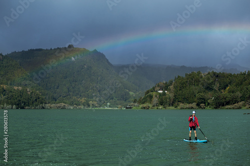A man in shorts and red jacket floats standing on a sapsurf on the lake in cloudy weather with beautiful rainbow background. Furnas Lake, Azores