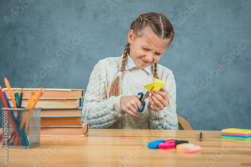 Pretty blond schollgirl is sitting at the desk and trying to cut the yellow paper. photo