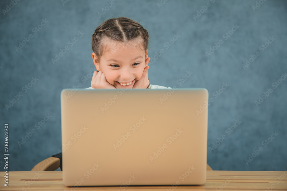 Cute little girl is looking at the screen of the computer with amusement and smiling widely. Cute little girl is looking at the screen of the computer with amusement and smiling widely.