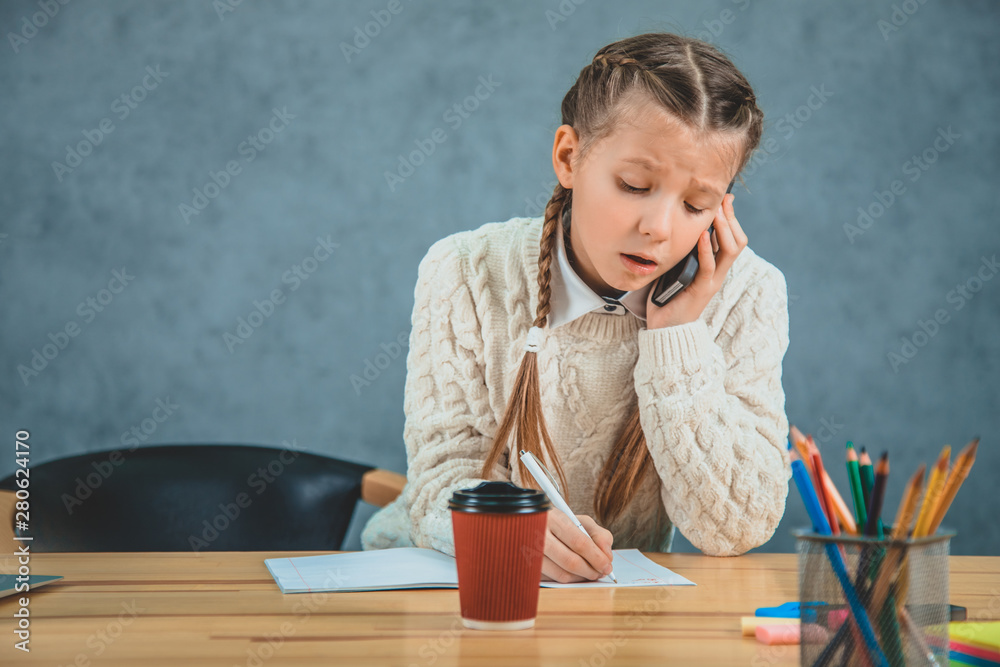 Serious schoolgirl in an uniform, with black cell near her ear a is concentrated on the work and talking. Time to create. Serious schoolgirl in an uniform, with black cell near her ear a is concentrated on the work and talking. Time to create.