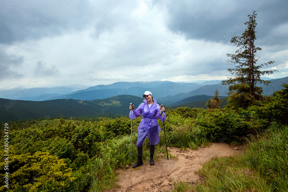 The girl, a tourist with Scandinavian sticks in a raincoat on the background of the beautiful Carpathian mountains. Rise in the mountains. Travel concept, leisure activity, vacation