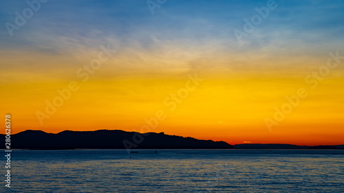 Early morning sunrise behind the mountains with sunlight reflection on the ocean/river. Landscape silhouette at dawn as the sun brings light to the sea coast. Start of the day with beautiful sunshine.