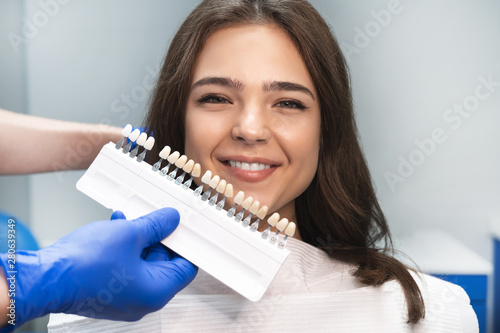 smiling happy brunette woman patient having appiontment in dental clinic picking up shade using tooth enamel scale held by dentist in blue gloves