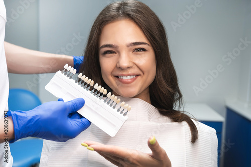 smiling brunette woman patient having appiontment in dental clinic picking up shade using tooth enamel scale held by dentist in blue gloves