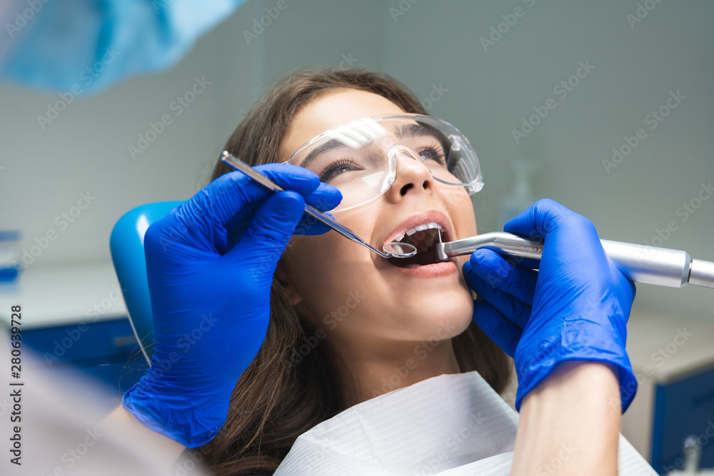 dentist in mask filling the patient's root canal while she is lying on dental chair wearing safety glasses under the medical lamp in dental office dentist in mask filling the patient's root canal while she is lying on dental chair wearing safety glasses under the medical lamp in dental office