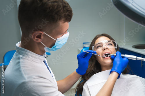 dentist in medical mask filling the patient's root canal while she is in dental chair wearing safety glasses under the medical lamp in clinic photo