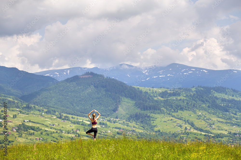 Attractive slim young woman doing yoga exercises outdoors on background of green mountains on sunny summer day.