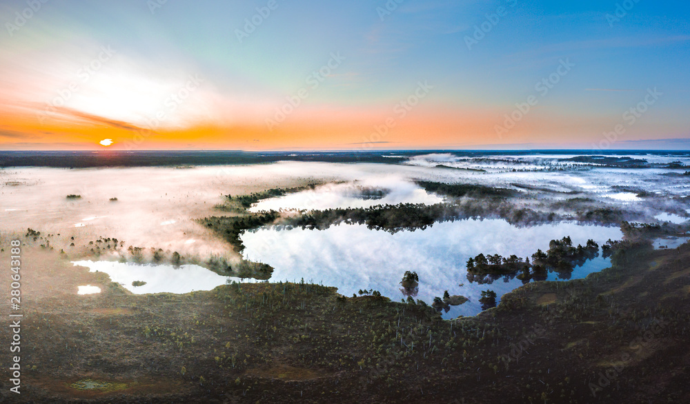 Warmly colored sunrise over a foggy swamp. Aerial view of stunning landscape at peat bog at Kemeri national park in Latvia. Wooden trail leading along the lake surrounded by pounds and forest. 