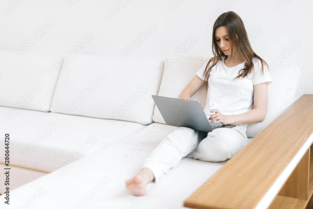 Young woman freelancer working at home on the white couch with laptop in light living room. Girl typing text on laptop. Female working on notebook at home in white interior Young woman freelancer working at home on the white couch with laptop in light living room. Girl typing text on laptop. Female working on notebook at home in white interior