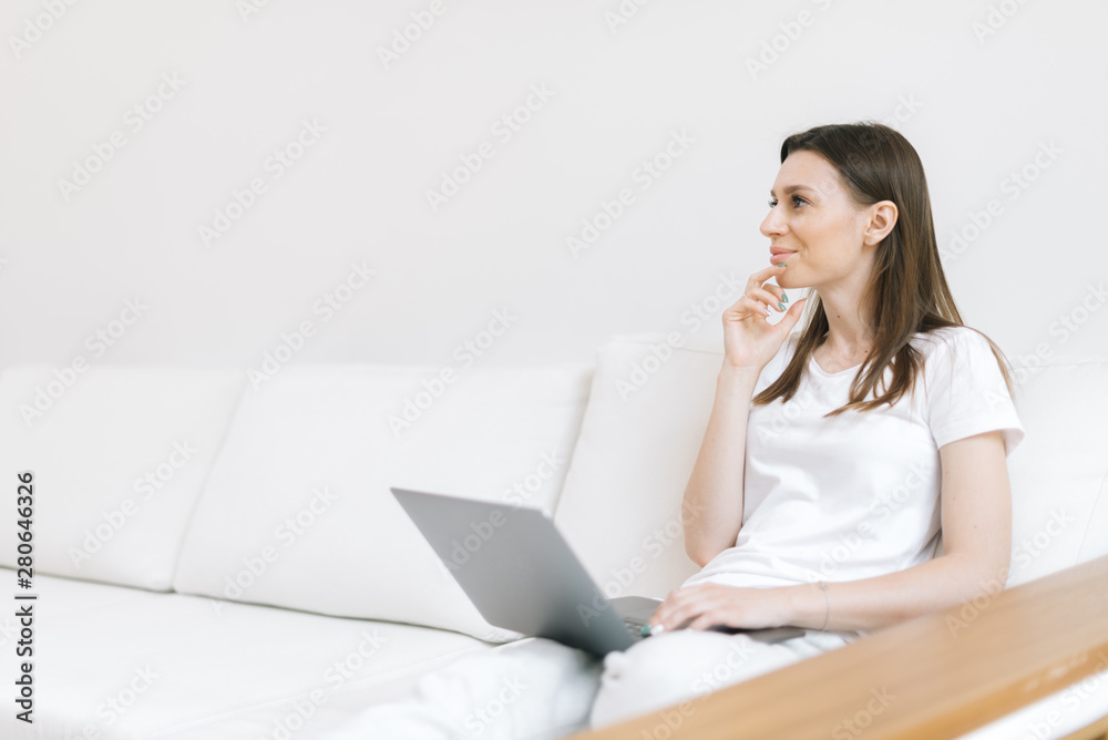 Smiling woman is sitting on white couch with laptop and thinking about something Smiling woman is sitting on white couch with laptop and thinking about something