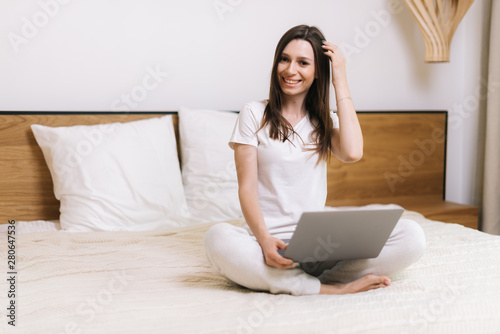 Happy smiling woman is sitting on bed, looking at camera and using laptop