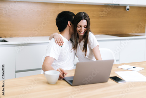 Young embracing couple is using laptop computer sitting on kitchen at home