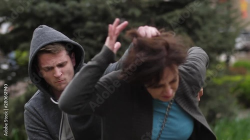 Close-up of adult female attacked from behind by arrogant thief grabbing hair and trying to steal purse during walk in city park. Brave woman applying Krav Maga martial arts to knock attaker out