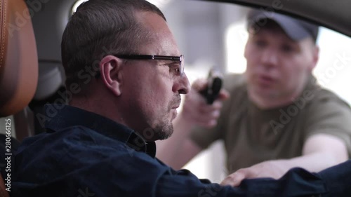 Close-up of aggressive mugger with firearm attacking male driving car and threatening with gun through open window. Brave driver grabbing gun and punching criminal intending to hijack car