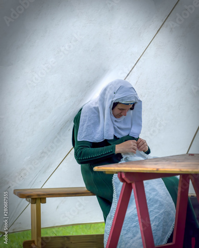 Woman in authentic medieval costume sat on a bench and table, sewing in a tent. The encampment at Cardiff Joust 2019.