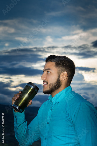 Profile portrait of a latin young man. He looks happy at the beginning of the day drinking his coffee.. The blue sky from dawn is at the back.