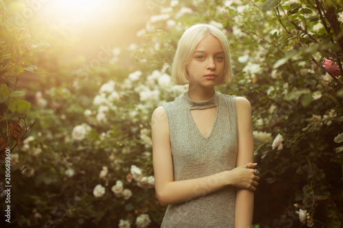 Portrait of beautiful young girl in white roses. Blond platinum girl with fair complexion. Sunset backlight. Image with selective focus and toning