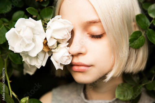 Portrait of beautiful young girl with closed eyes in white roses. Blond platinum girl with fair complexion and roses in front of her face