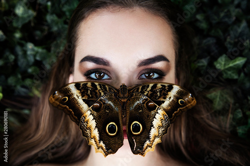 Portrait of beautiful young girl with butterfly sitting on her face. Big black butterfly Papilio lowi sat on the woman's forehead. Symmetrical position.