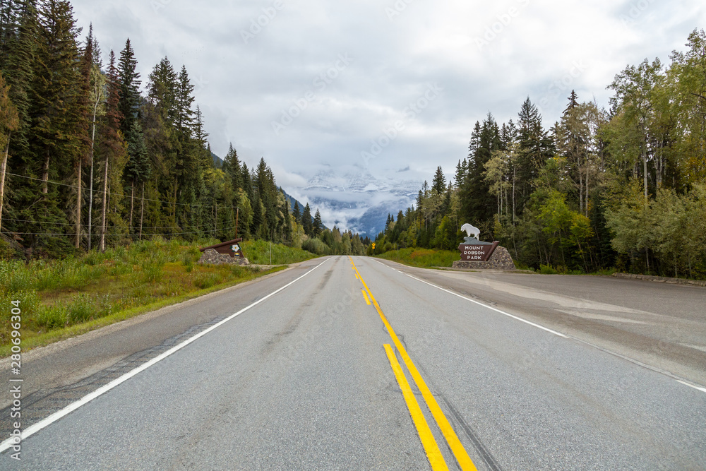 Fototapeta premium Road into the Canadian Rockies with Mount Robson in the background covered with fog and the Mount Robson Provincial Park sign beside the road