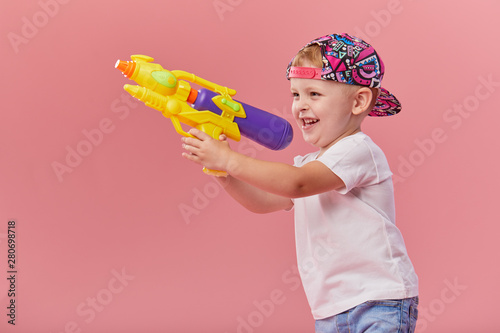 Happy little toddler boy  in summer clothes holds toy water gun isolated on pink wall background. Children studio portrait. People childhood lifestyle concept.