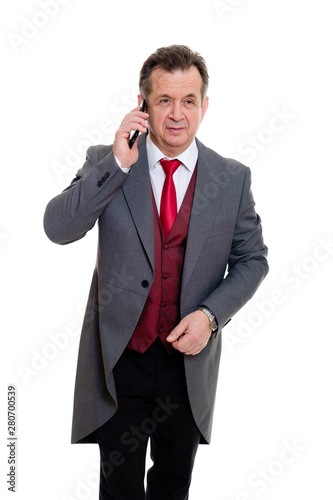 Portrait of handsome confident caucasian mature businessman 55-60 years old in grey tuxedo and red vest. Rich senior man talk by phone and looking to camera on isolated white background