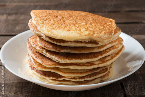 Heap of homemade pancakes without cream lies on round white ceramic plate and old rustic wooden table