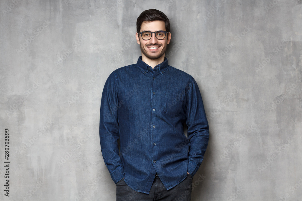 Young handsome man wering eyeglasses, smiling and feeling confident, standing against gray textured wall background